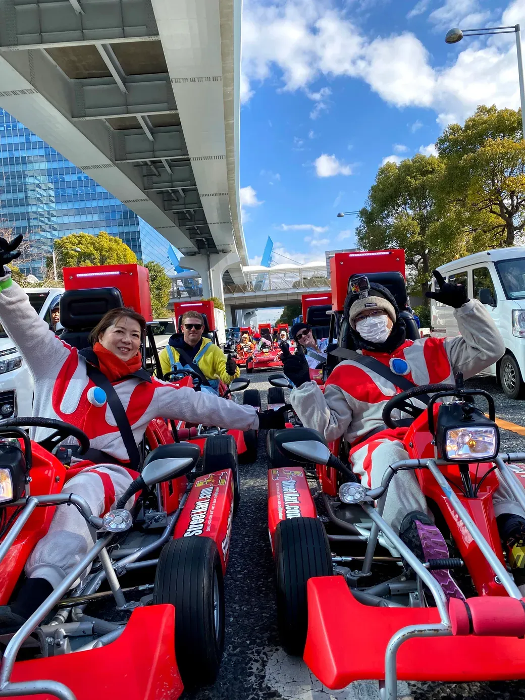 People in red and white racing outfits in red go-karts wave at the camera under a highway overpass on a sunny day.