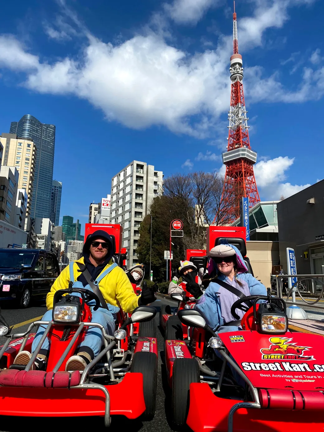 People riding bright red go-karts down a city street, with Tokyo Tower visible in the distance against a blue sky.