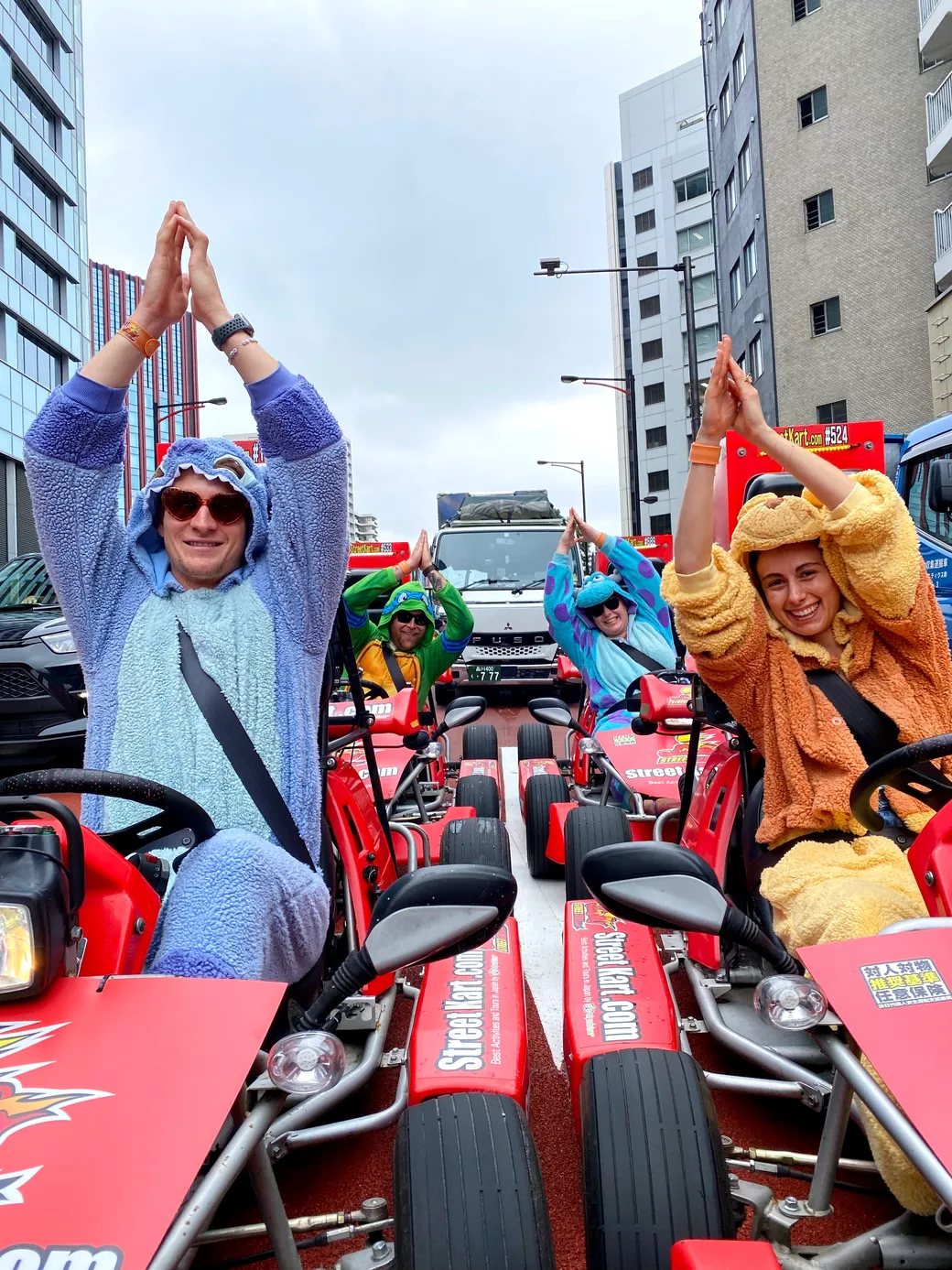 People in fuzzy animal onesies pose in red go-karts on a city street, hands raised together above their heads in a playful gesture.